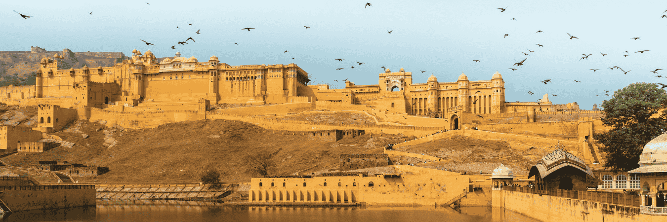 Golden-hued Amber Fort perched on Aravalli hills above Maota Lake in Jaipur, Rajasthan, with birds flying and reflections in the water under a clear sky