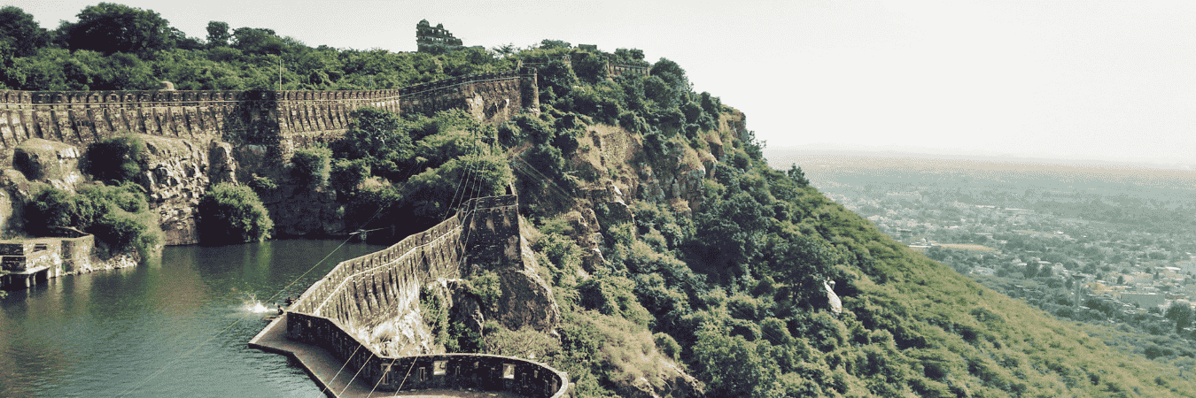 Panoramic view of an ancient stone fort wall running along a lush green hillside above a calm lake, with the cityscape visible in the distant background