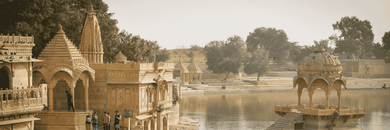 Scenic view of intricately carved golden sandstone temples and chhatris reflecting in a calm lake at Jaisalmer, with wide ghats, trees, and soft evening light in the background.